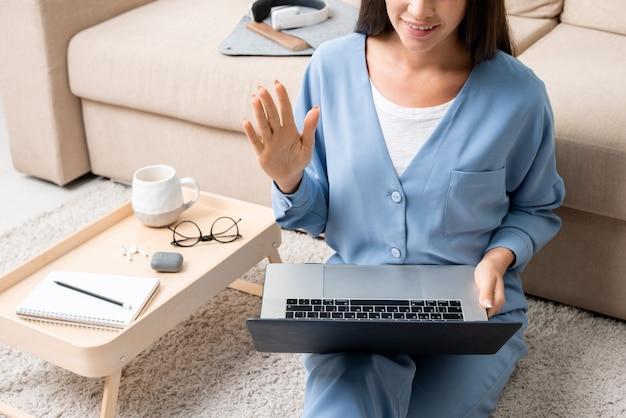 Фото Молодая современная женщина в синей пижаме машет рукой во время онлайн-общения со своими коллегами, сидя на полу дома